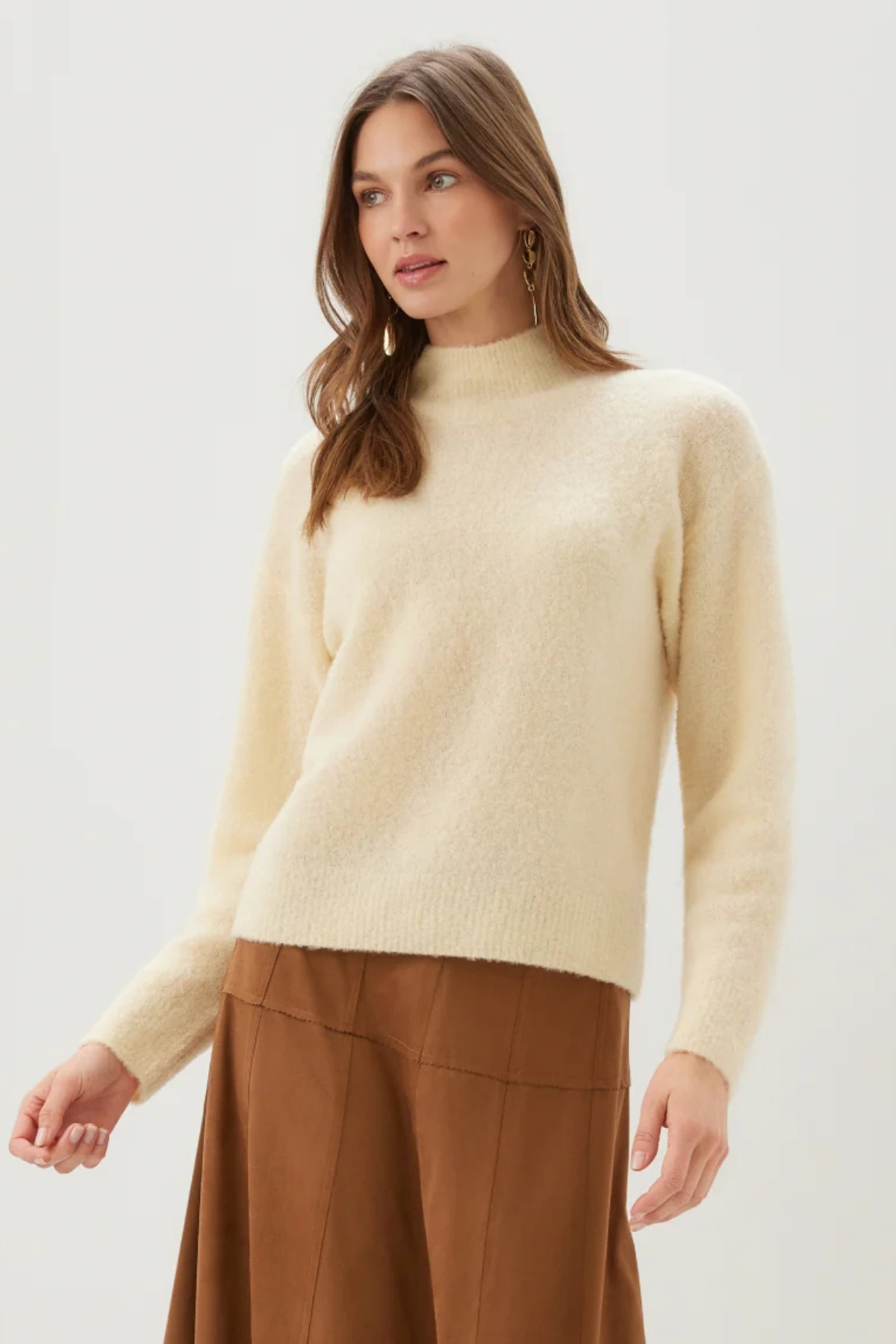 A woman wearing a cream-colored, fuzzy mock-neck knit sweater and brown wide-leg trousers, photographed against a clean, light gray studio background.