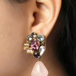A close-up of a model's ear wearing a statement dangle earring. The earring features a cluster of colorful rhinestones in pink, purple, blue, and gold-tone metal, with a dangling light pink rose quartz teardrop charm.