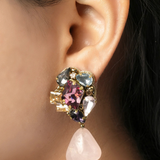 A close-up of a model's ear wearing a statement dangle earring. The earring features a cluster of colorful rhinestones in pink, purple, blue, and gold-tone metal, with a dangling light pink rose quartz teardrop charm.