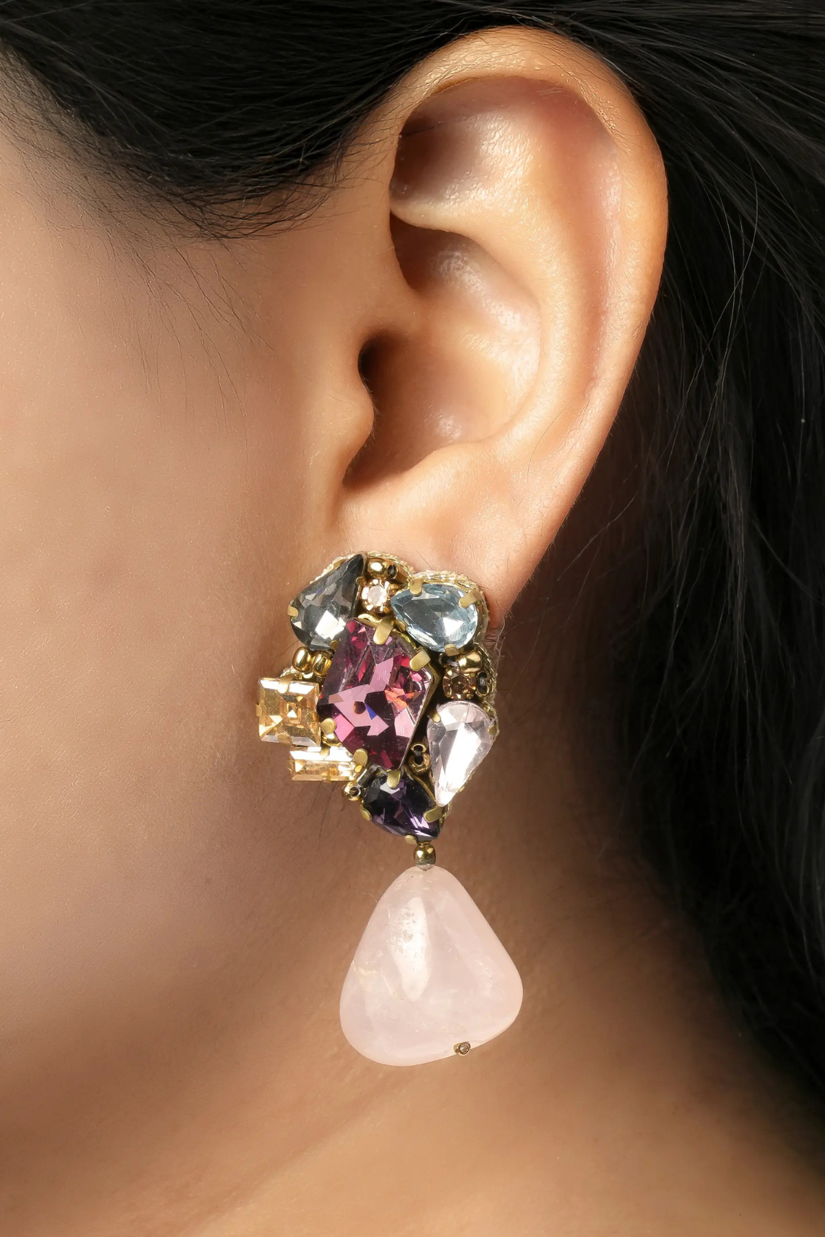 A close-up of a model's ear wearing a statement dangle earring. The earring features a cluster of colorful rhinestones in pink, purple, blue, and gold-tone metal, with a dangling light pink rose quartz teardrop charm.
