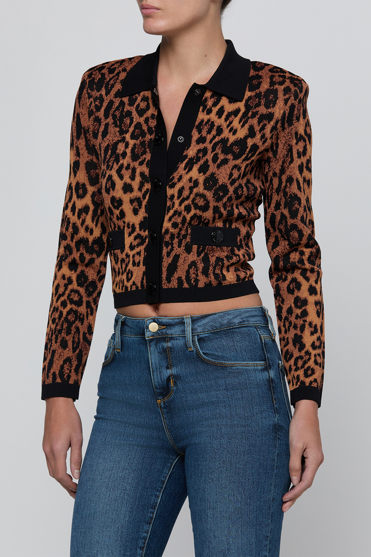 A cropped long-sleeve knit cardigan with a brown and black leopard print, black collar, and black button placket, worn by a model with blue jeans against a light gray background.