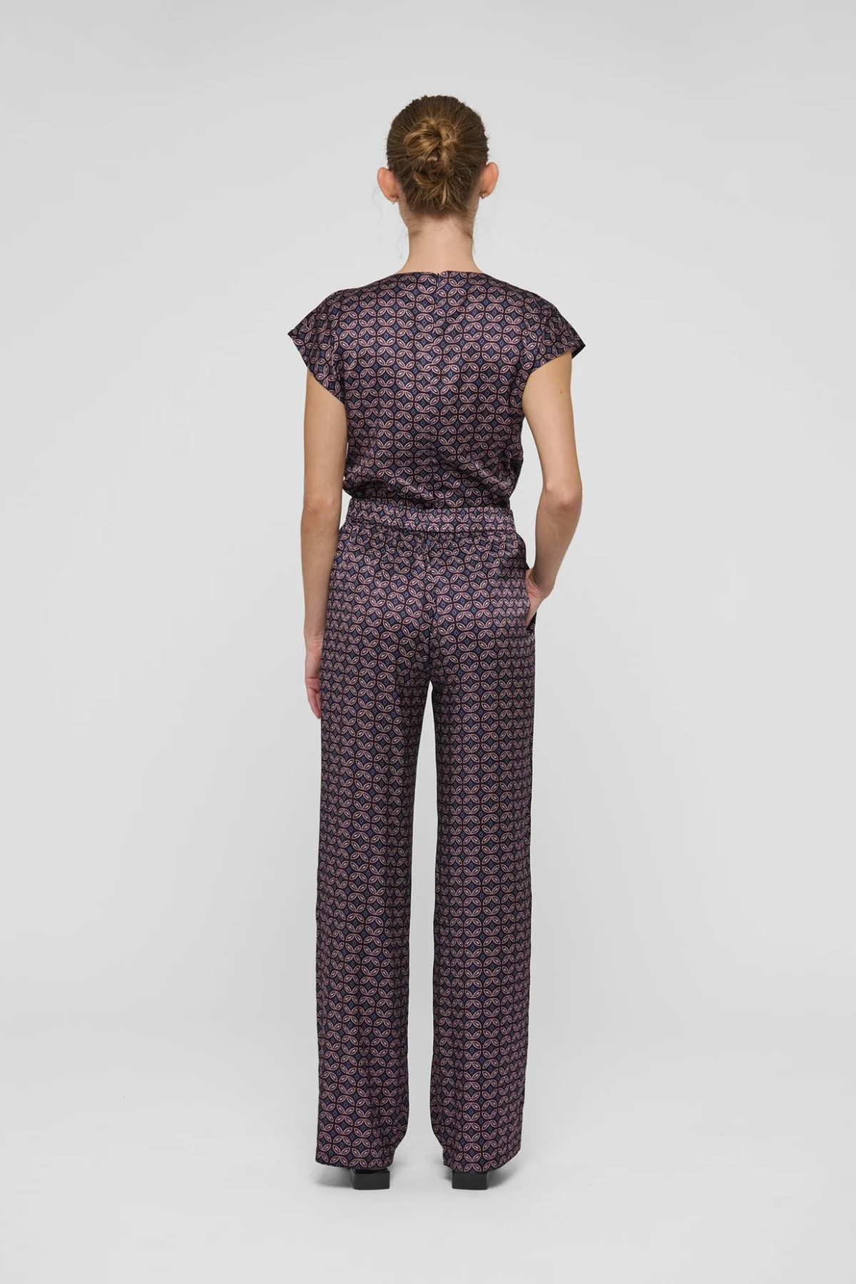 A model viewed from the back wearing a matching short-sleeve top and wide-leg trousers set in a dark purple and black geometric pattern, set against a plain light gray background.