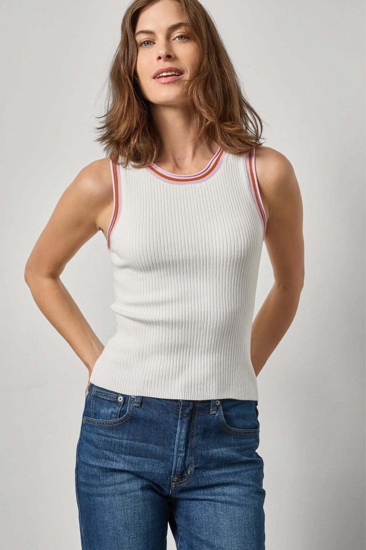 Woman wearing a white ribbed tank top with red and orange trim and blue jeans on a light gray background