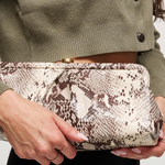 A close-up of a person holding a snakeskin-patterned clutch bag. The person is wearing an olive green cropped cardigan and black pants, with manicured nails visible on the hands.