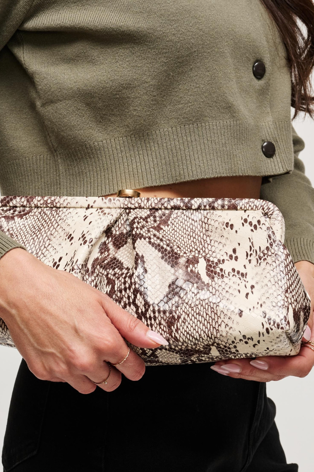 A close-up of a person holding a snakeskin-patterned clutch bag. The person is wearing an olive green cropped cardigan and black pants, with manicured nails visible on the hands.