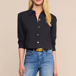 A blonde model wearing a black long-sleeve button-up shirt with gold buttons and blue jeans, standing against a solid beige background.