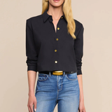 A blonde model wearing a black long-sleeve button-up shirt with gold buttons and blue jeans, standing against a solid beige background.