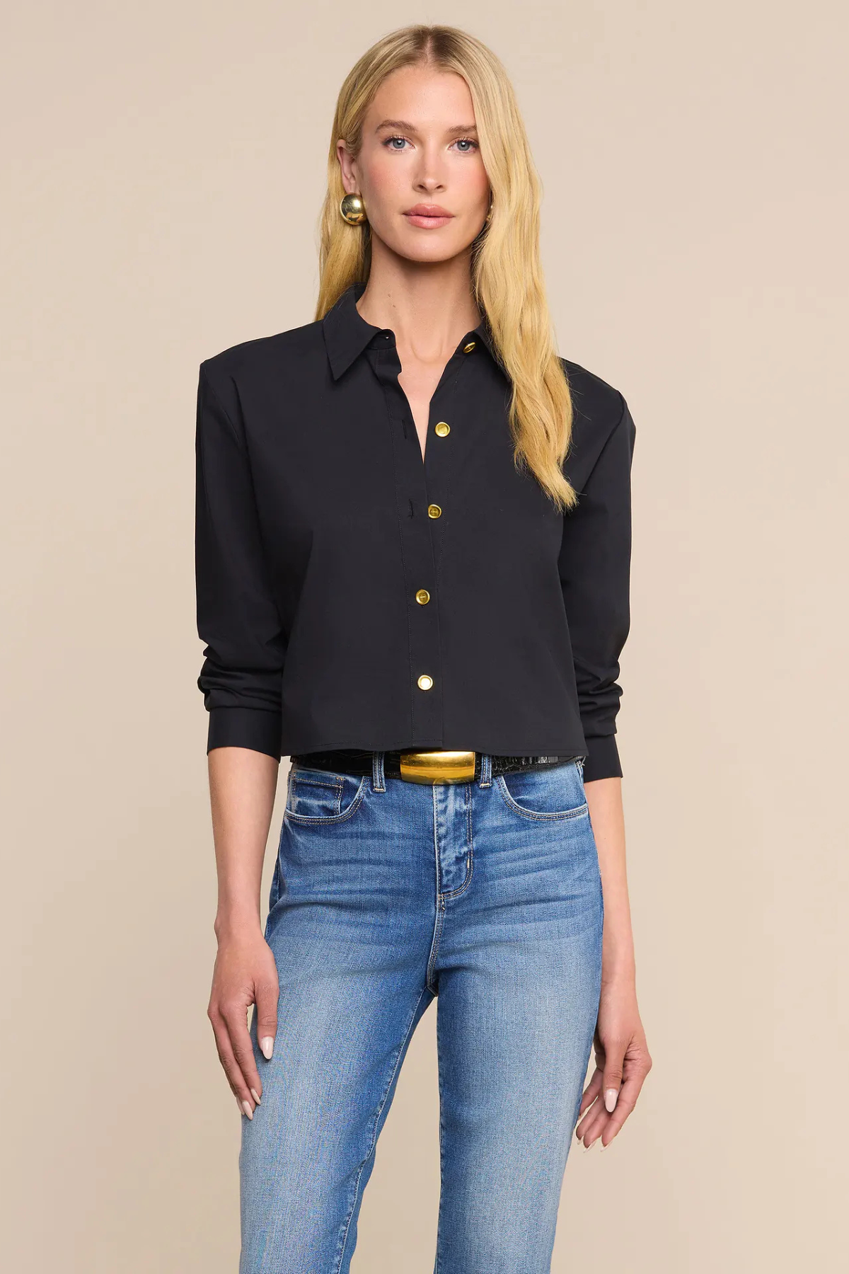 A blonde model wearing a black long-sleeve button-up shirt with gold buttons and blue jeans, standing against a solid beige background.