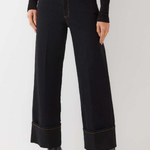 A model wearing a pair of black wide-leg cropped jeans with rolled cuffs and gold contrast stitching, paired with a black long-sleeve top and black boots, against a plain light gray studio background.
