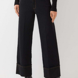 A model wearing a pair of black wide-leg cropped jeans with rolled cuffs and gold contrast stitching, paired with a black long-sleeve top and black boots, against a plain light gray studio background.