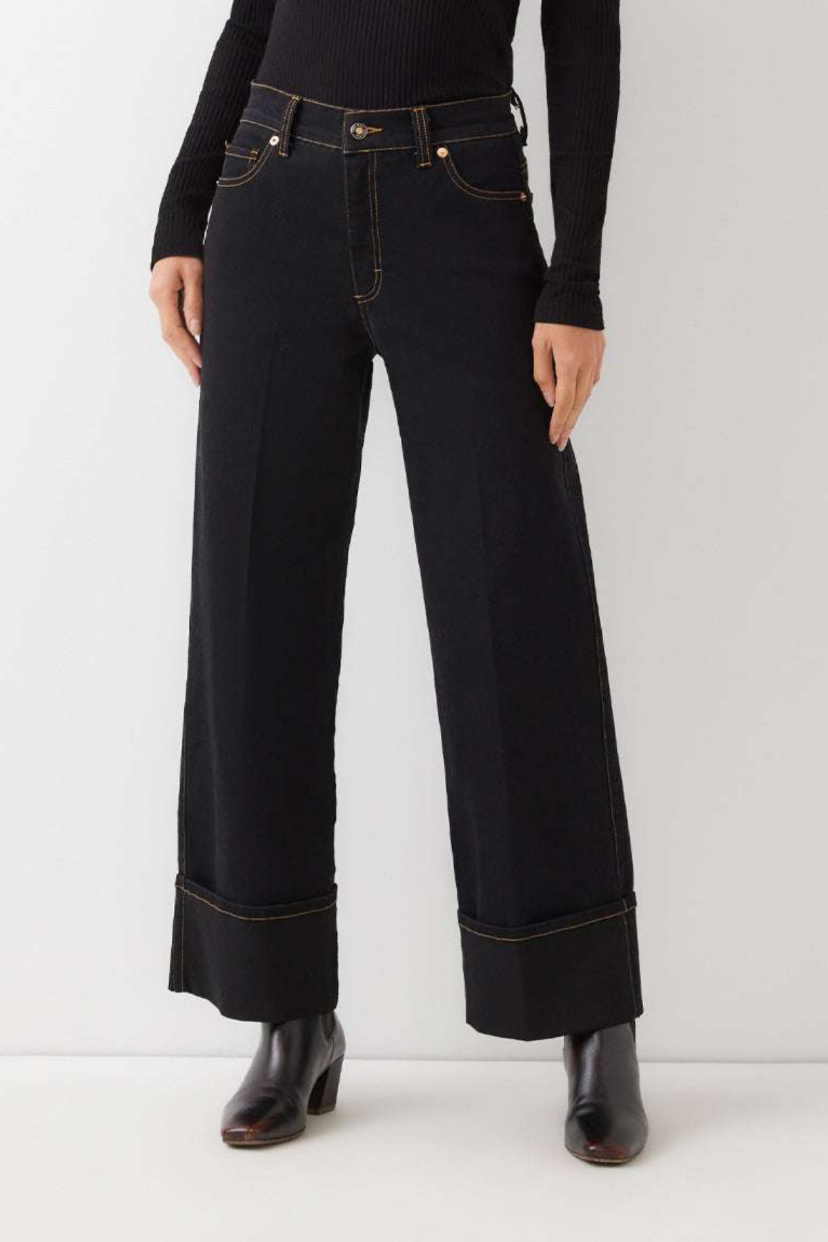 A model wearing a pair of black wide-leg cropped jeans with rolled cuffs and gold contrast stitching, paired with a black long-sleeve top and black boots, against a plain light gray studio background.