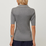 A woman viewed from the back wearing a gray short-sleeve ribbed knit top with a decorative silver beaded collar, paired with navy pants, against a light gray studio background.