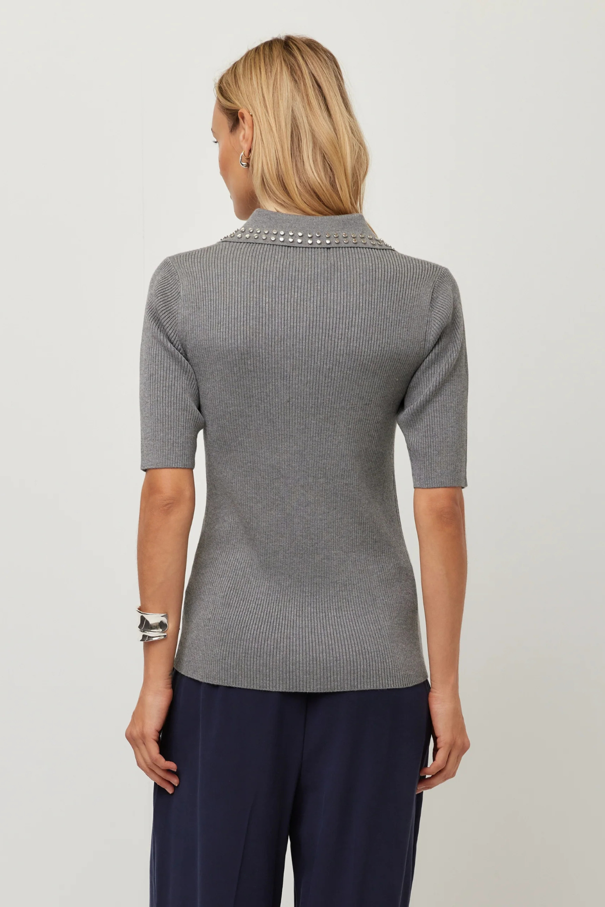 A woman viewed from the back wearing a gray short-sleeve ribbed knit top with a decorative silver beaded collar, paired with navy pants, against a light gray studio background.