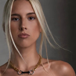 A close-up portrait of a blonde woman wearing a gold chain necklace with a central black bar and silver-toned rings, styled with a black strapless top against a dark gray background.