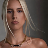 A close-up portrait of a blonde woman wearing a gold chain necklace with a central black bar and silver-toned rings, styled with a black strapless top against a dark gray background.