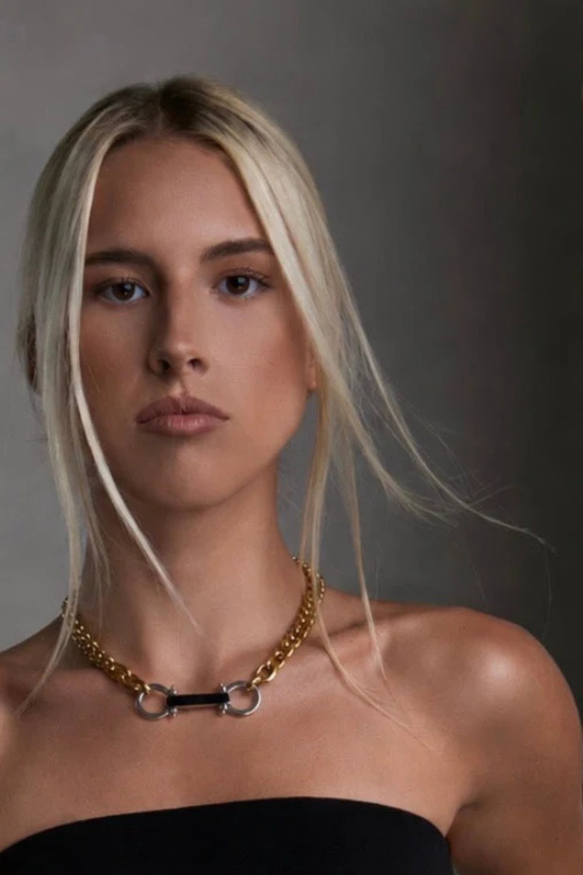 A close-up portrait of a blonde woman wearing a gold chain necklace with a central black bar and silver-toned rings, styled with a black strapless top against a dark gray background.
