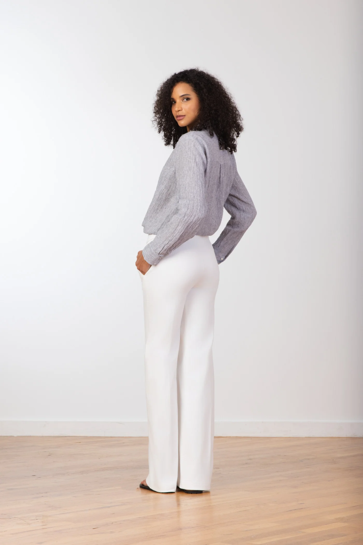 Woman wearing a gray sweater and white pants standing against a plain white background