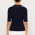 A woman shown from the back wearing a navy blue short-sleeve knit top with a studded collar, paired with white pants, against a plain light gray studio background.