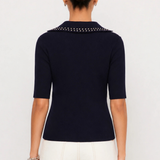 A woman shown from the back wearing a navy blue short-sleeve knit top with a studded collar, paired with white pants, against a plain light gray studio background.