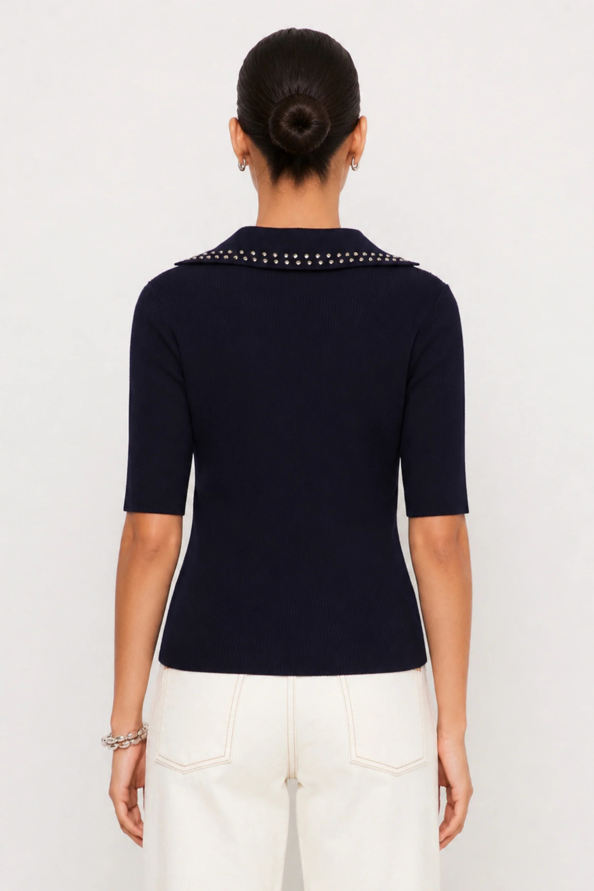 A woman shown from the back wearing a navy blue short-sleeve knit top with a studded collar, paired with white pants, against a plain light gray studio background.