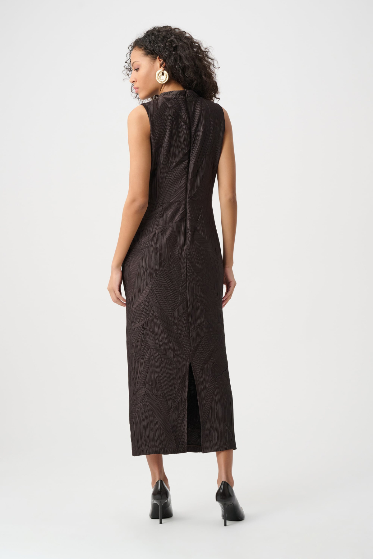 A woman shown from the back wearing a sleeveless dark brown midi dress with a textured crinkled fabric and a front slit, paired with black pointed-toe heels against a light gray studio background.