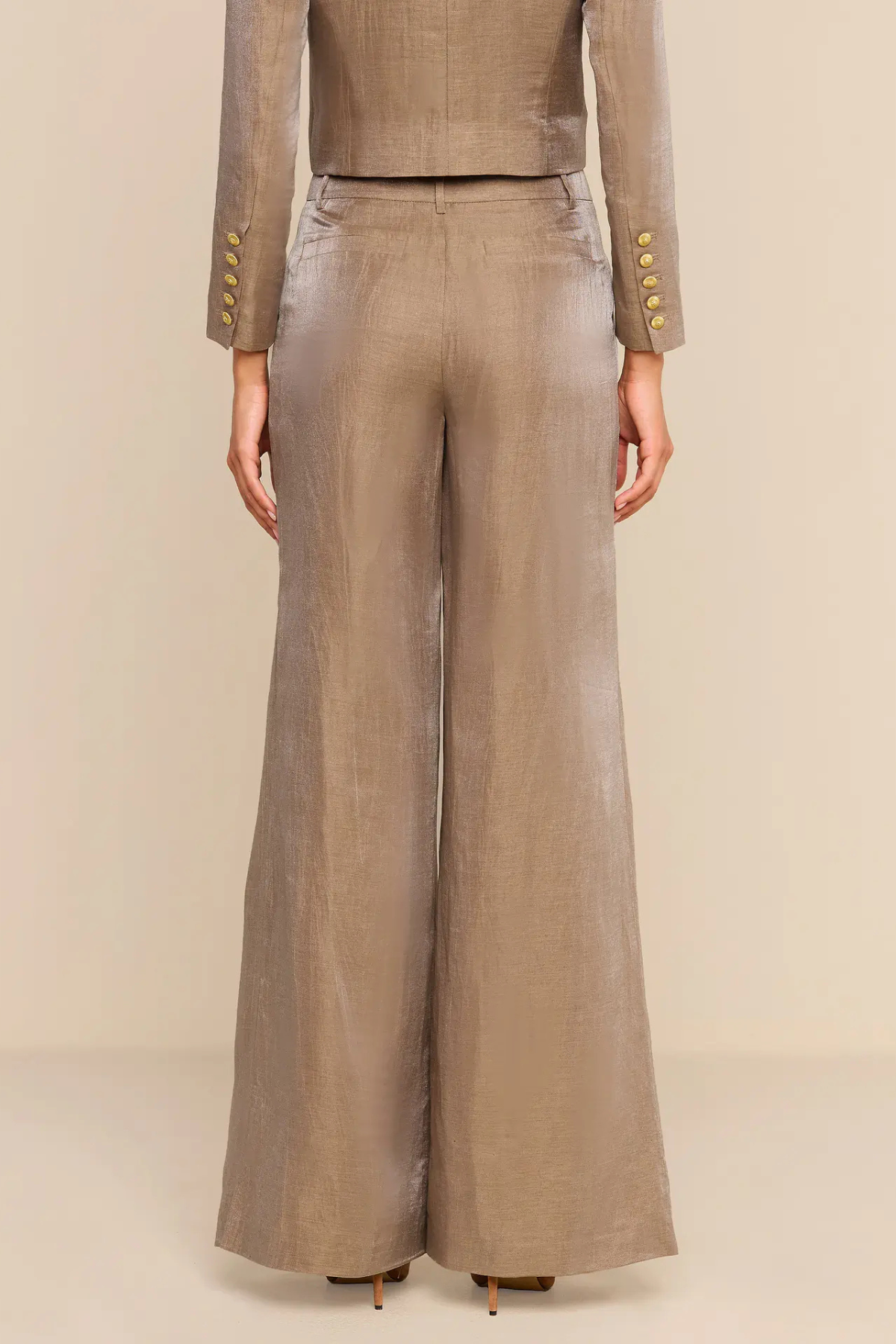 A model shown from the back wearing a matching taupe-beige two-piece set with wide-leg trousers and a cropped long-sleeve top, both featuring gold button accents on the cuffs.