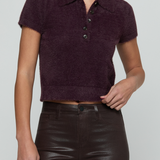 A model wearing a short-sleeve fuzzy knit polo shirt in deep plum purple, paired with dark brown faux-leather pants, against a solid light gray background.