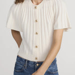 Woman wearing a white ribbed blouse with gold buttons and blue jeans on a plain background