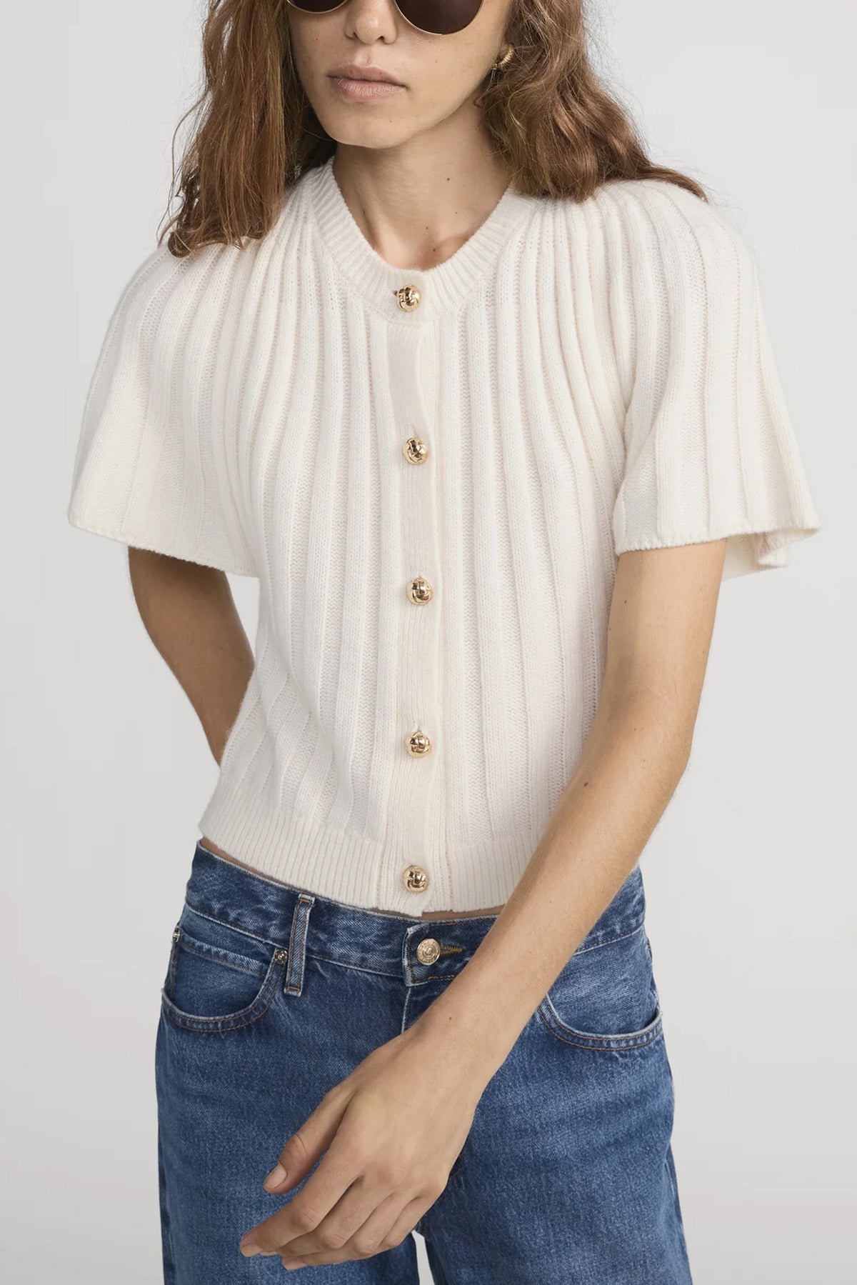 Woman wearing a white ribbed blouse with gold buttons and blue jeans on a plain background