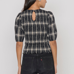A woman shown from the back wearing a black and beige plaid puff-sleeve top with a keyhole cutout at the neck, paired with dark jeans, against a plain light gray studio background.
