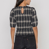 A woman shown from the back wearing a black and beige plaid puff-sleeve top with a keyhole cutout at the neck, paired with dark jeans, against a plain light gray studio background.