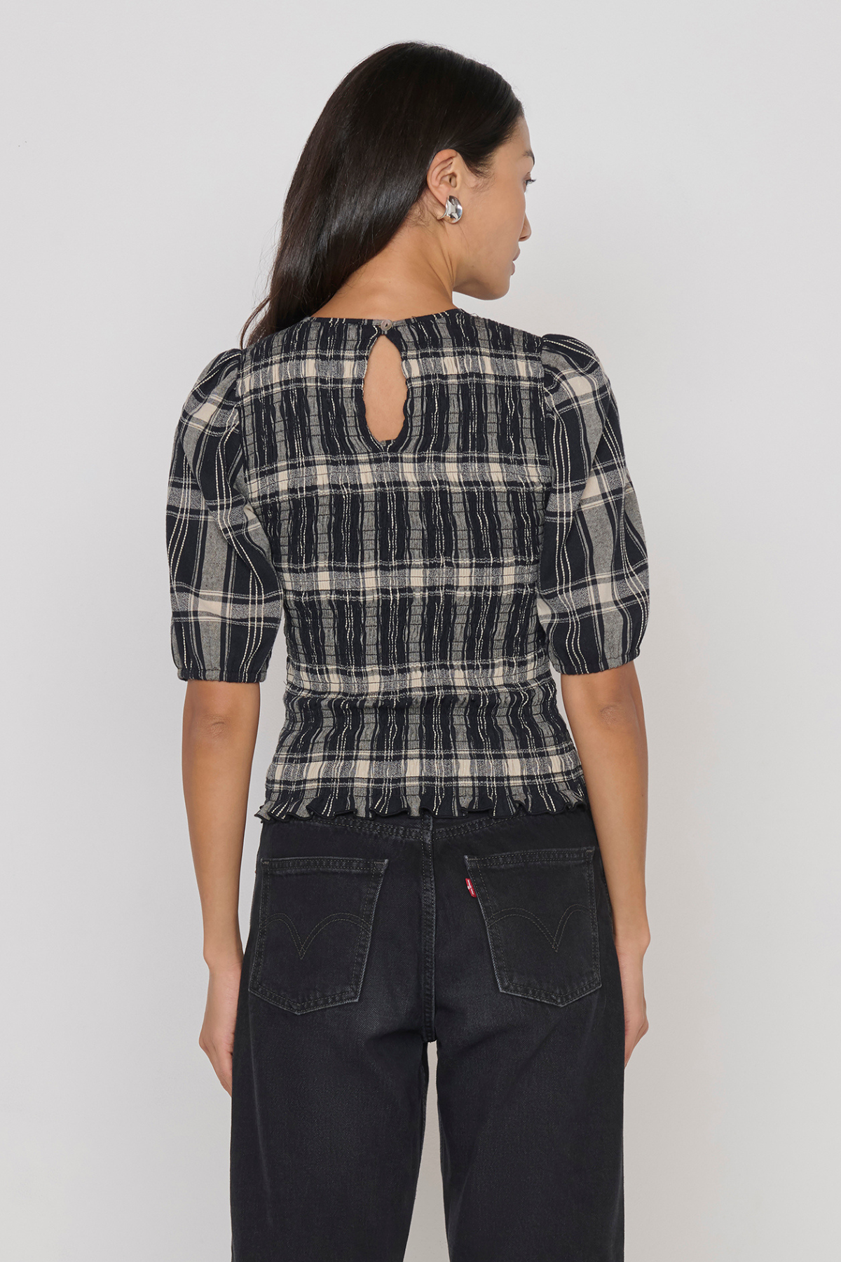 A woman shown from the back wearing a black and beige plaid puff-sleeve top with a keyhole cutout at the neck, paired with dark jeans, against a plain light gray studio background.