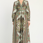 A woman modeling a long-sleeved midi dress with a detailed botanical and tribal pattern in olive green, brown, beige, and rust red, set against a plain light gray studio background.