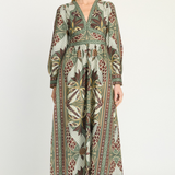 A woman modeling a long-sleeved midi dress with a detailed botanical and tribal pattern in olive green, brown, beige, and rust red, set against a plain light gray studio background.