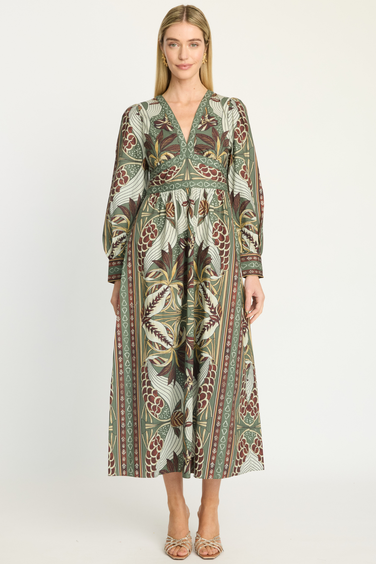A woman modeling a long-sleeved midi dress with a detailed botanical and tribal pattern in olive green, brown, beige, and rust red, set against a plain light gray studio background.