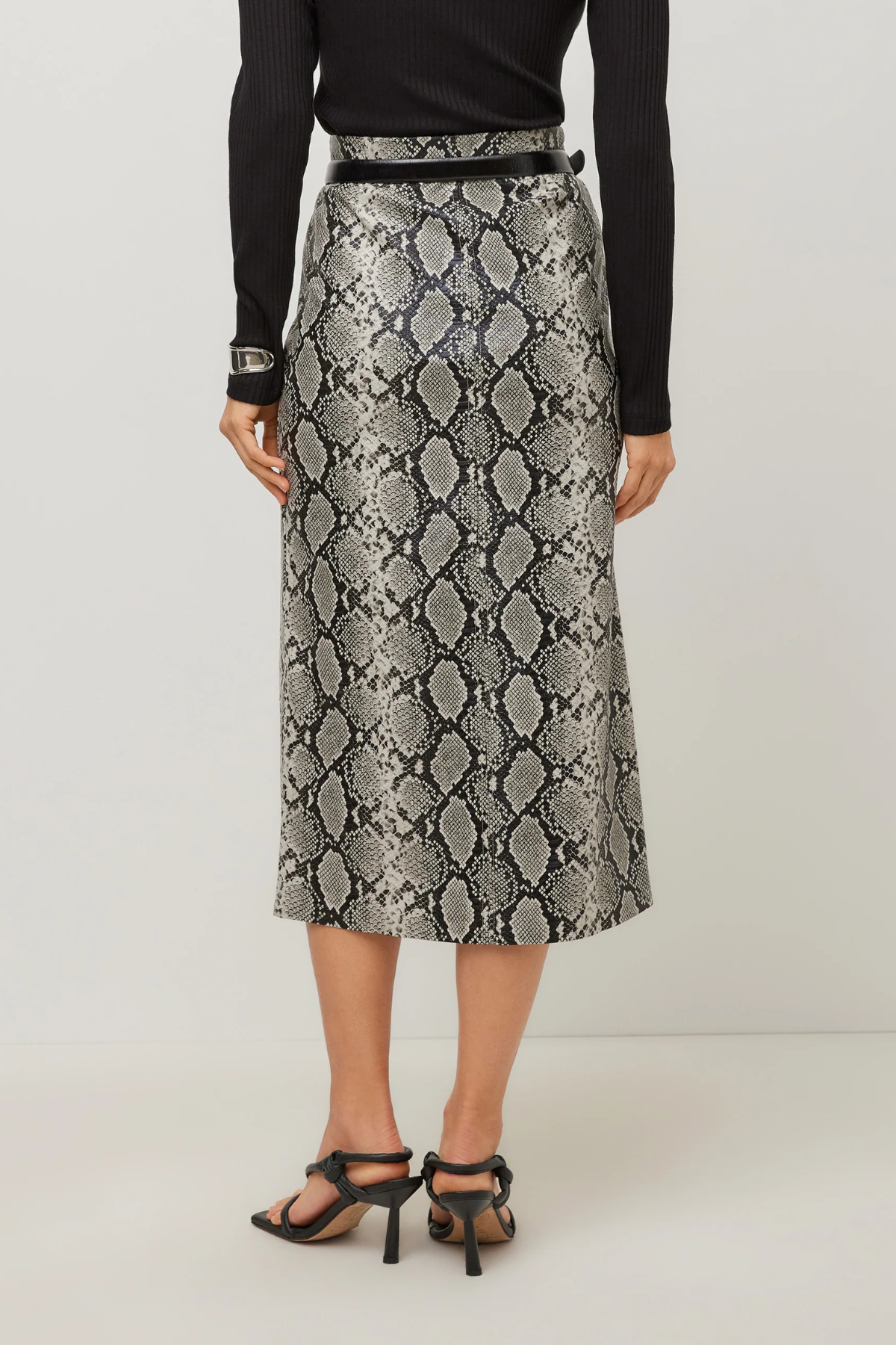 A model from the back wearing a high-waisted midi skirt with a gray and black snakeskin pattern, styled with a black top, black belt, and black strappy heels against a light gray studio background.