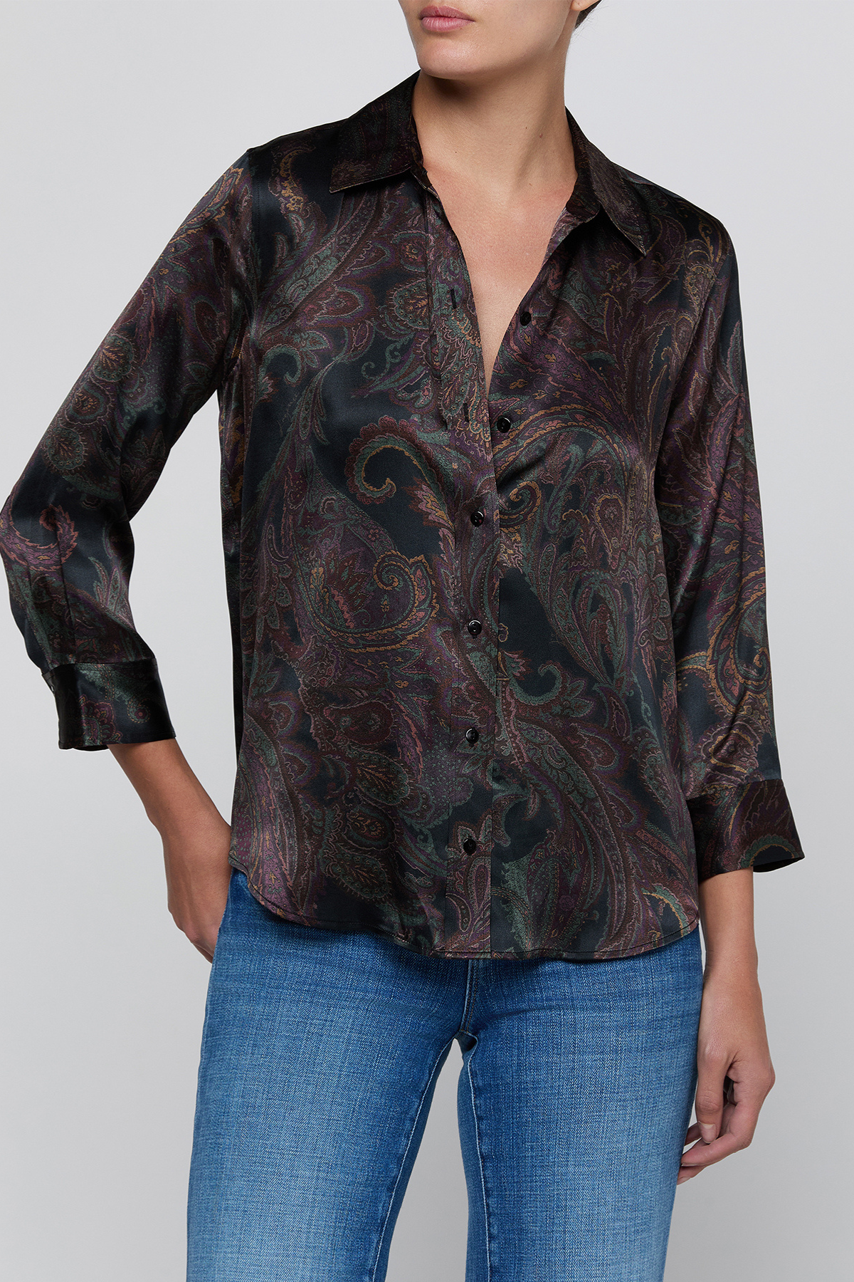 A woman wearing a long-sleeve button-up shirt with a dark paisley pattern in black, brown, and muted green, paired with blue jeans, against a plain light gray background.
