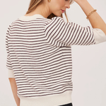 A woman wearing a cream and dark brown horizontally striped knit sweater, shown from a back-three-quarter angle against a plain light background.
