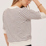 A woman wearing a cream and dark brown horizontally striped knit sweater, shown from a back-three-quarter angle against a plain light background.