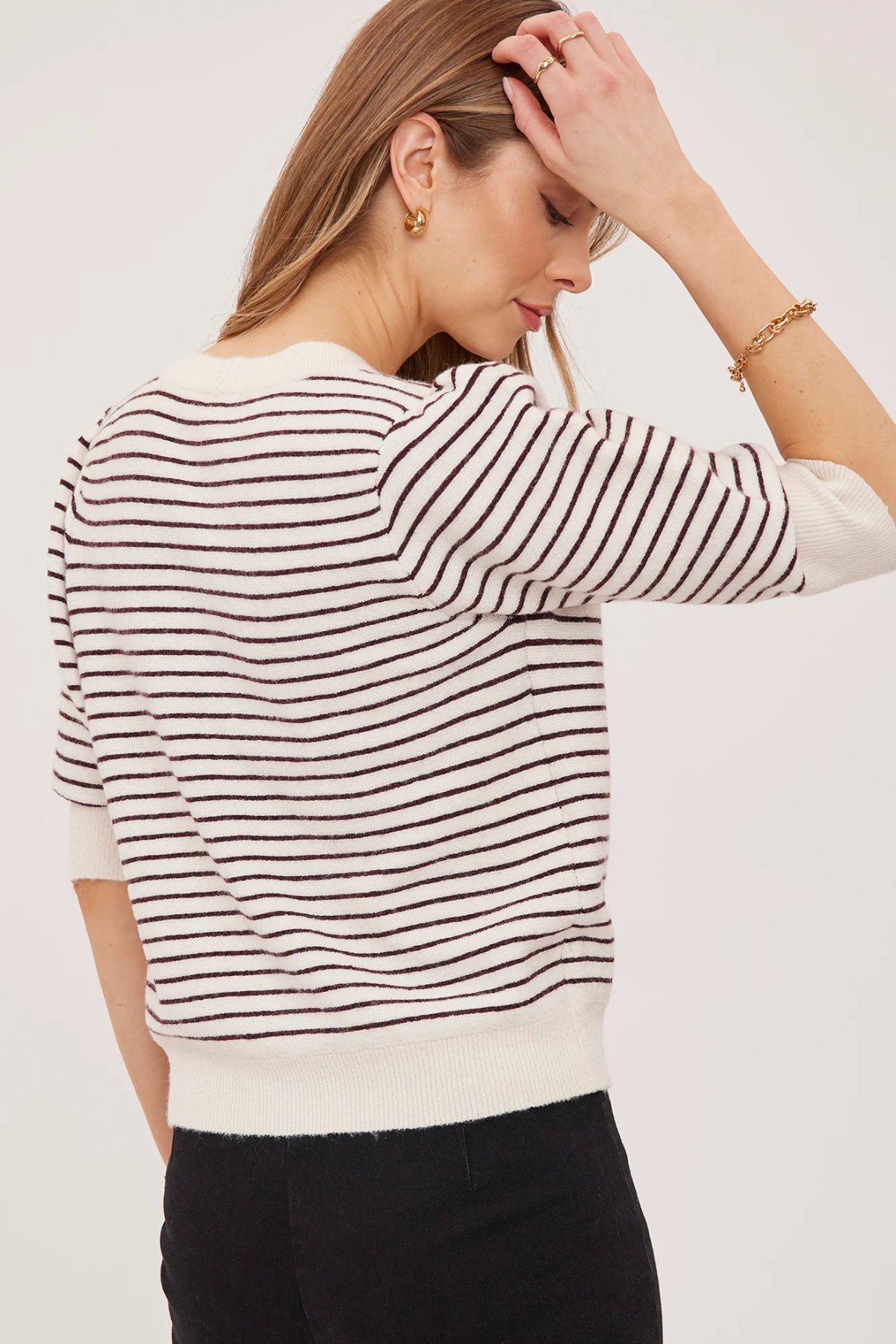 A woman wearing a cream and dark brown horizontally striped knit sweater, shown from a back-three-quarter angle against a plain light background.