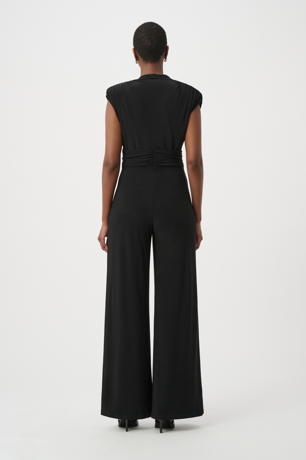 A model viewed from the back wearing a black sleeveless jumpsuit with a gathered waist and wide legs, set against a plain light gray studio background.