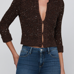 A woman wearing a dark brown cropped cardigan with sequins and blue jeans, shown from the mouth down against a light gray studio background.
