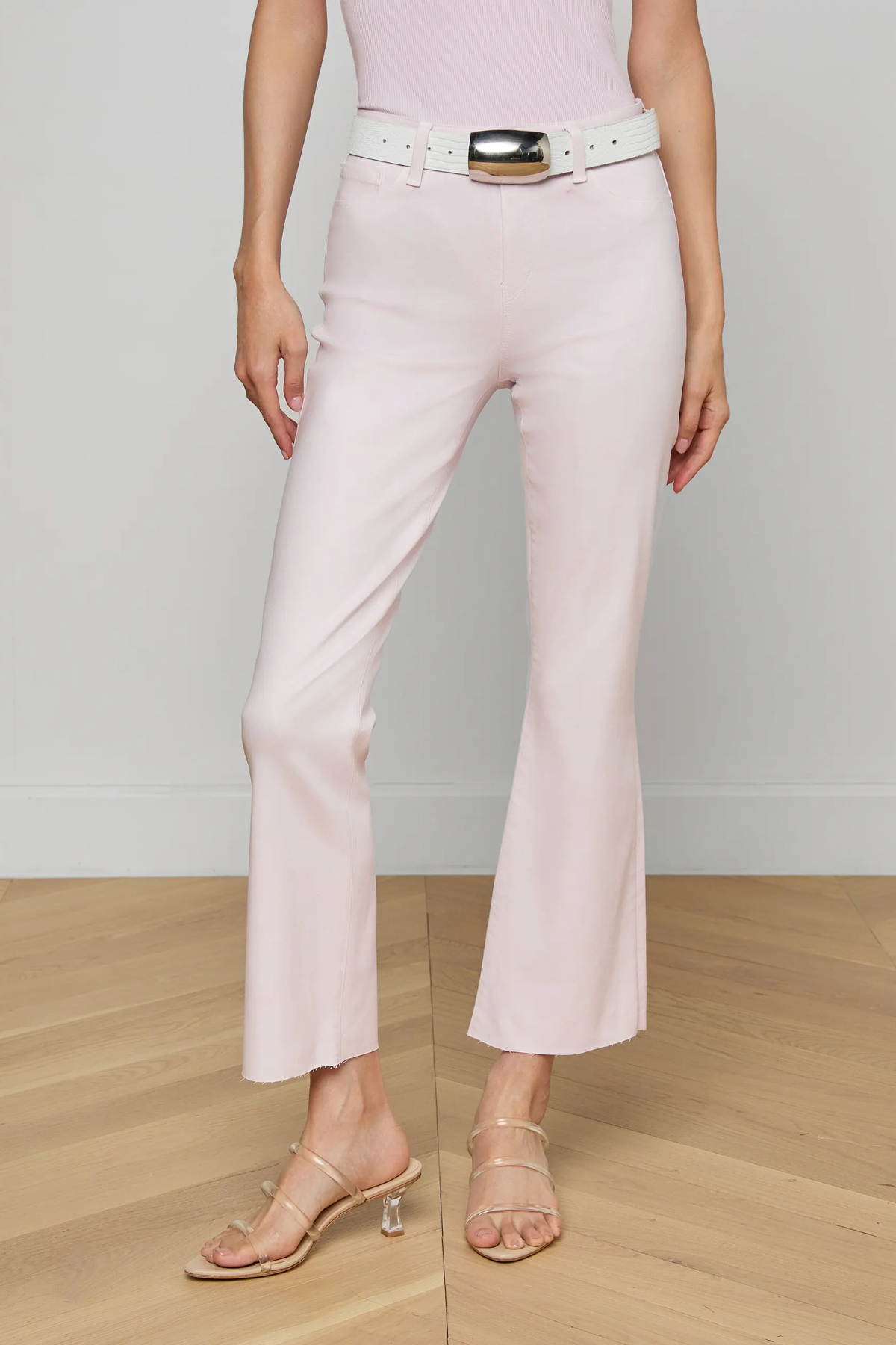 Person wearing light pink pants with a white belt and clear high-heeled sandals on a wooden floor.