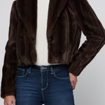A model wearing a dark brown faux fur cropped jacket over a cream turtleneck and blue jeans, shown from the neck down against a light gray studio background.