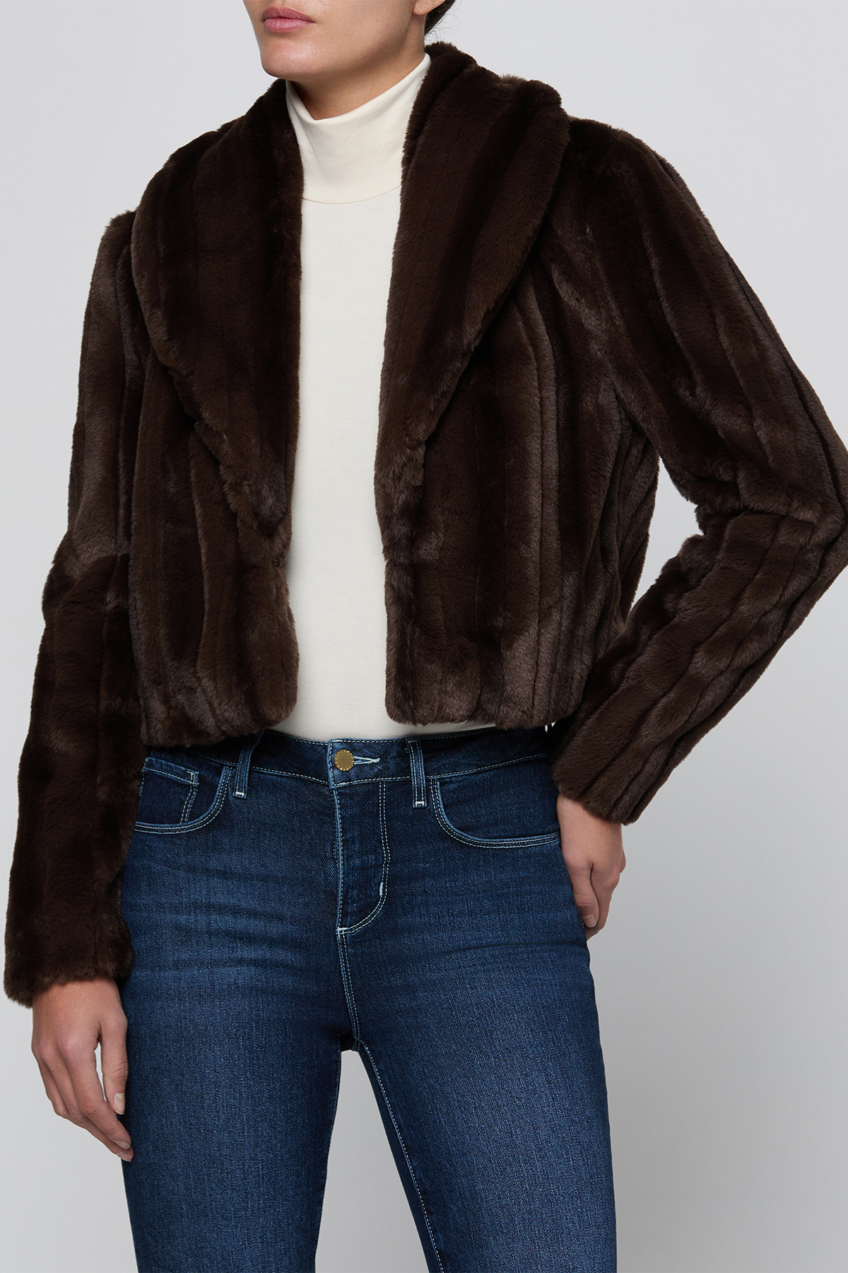 A model wearing a dark brown faux fur cropped jacket over a cream turtleneck and blue jeans, shown from the neck down against a light gray studio background.