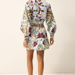 A woman shown from the back wearing a long-sleeve mini dress with a colorful floral and botanical print on a light background, paired with tan strappy flat sandals, against a plain off-white studio backdrop.