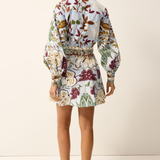 A woman shown from the back wearing a long-sleeve mini dress with a colorful floral and botanical print on a light background, paired with tan strappy flat sandals, against a plain off-white studio backdrop.