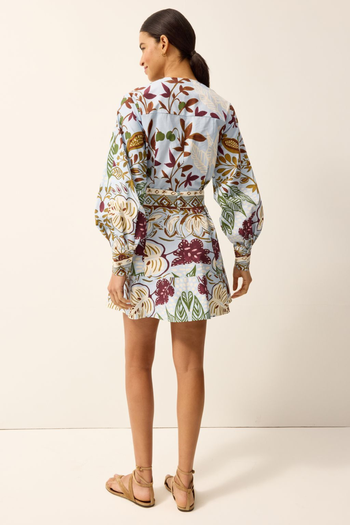 A woman shown from the back wearing a long-sleeve mini dress with a colorful floral and botanical print on a light background, paired with tan strappy flat sandals, against a plain off-white studio backdrop.