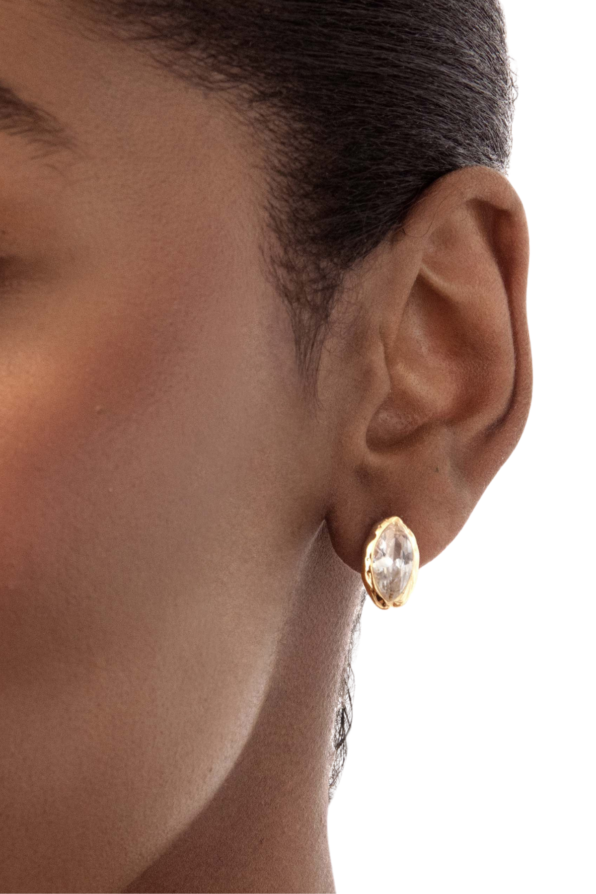 A close-up of a model's ear wearing a small gold stud earring with a marquise-cut clear gemstone.