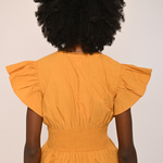 A person shown from the back wearing a mustard yellow peplum top with ruffled short sleeves and a smocked waist, paired with dark brown pants, against a plain off-white background.